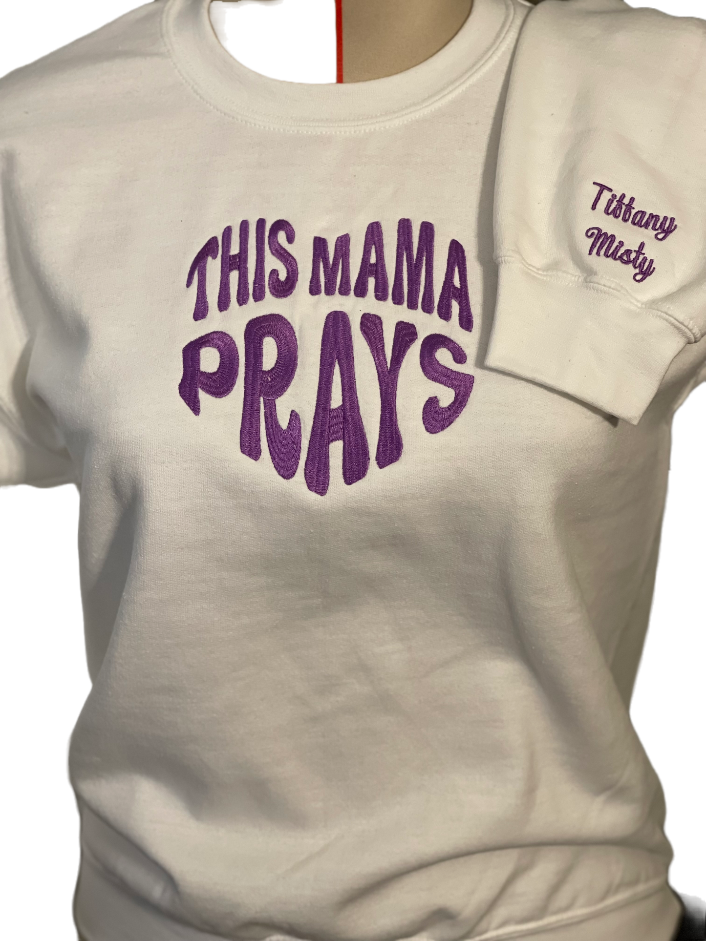 white long sleeve sweatshirt, with embroidered words "THIS MAMA PRAYS" the letters and making a shape of a heart, the thread in the picture is purple but there are choices of different colors to choose from. the lower sleeve have names Tiffany and Misty, the children's name of the Mama. Thread is matching purple color as the main word in the heart shape in the center of the shirt front. Personalize the names on the sleeves. up to 4 names on each sleeve.