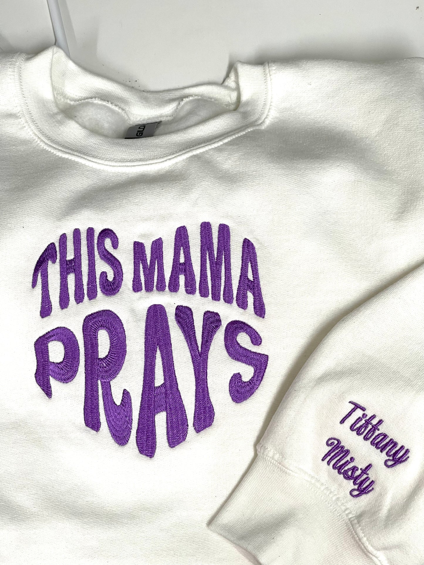 white long sleeve sweatshirt, with embroidered words "THIS MAMA PRAYS" the letters and making a shape of a heart, the thread in the picture is purple but there are choices of different colors to choose from. the lower sleeve have names Tiffany and Misty, the children's name of the Mama. Thread is matching purple color as the main word in the heart shape in the center of the shirt front. Personalize the names on the sleeves. up to 4 names on each sleeve.